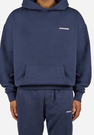 Mann trägt dunkelblauen Kapuzenpullover und passende Jogginghose, beide mit kleinen weißen "PEGADOR" Logos auf Brust und Oberschenkel.