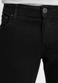 Černé džíny z denimu s klasickým designem, které mají přední zapínání na knoflíky, standardní kapsy a hladkou texturu bez viditelných vzorů.