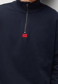 Felpa blu navy con colletto alto, zip frontale a metà e etichetta logo rettangolare rossa. Texture morbida e design casual.