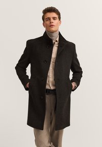 Czarnoszary płaszcz z mieszanki wełny z wysokim kołnierzem, zapinany na guziki, z dwiema bocznymi kieszeniami i dopasowanym krojem, noszony na jasnej beżowej koszuli.