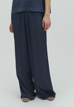 Persona che indossa pantaloni larghi blu scuro e scarpe beige, in piedi davanti a uno sfondo chiaro e semplice.