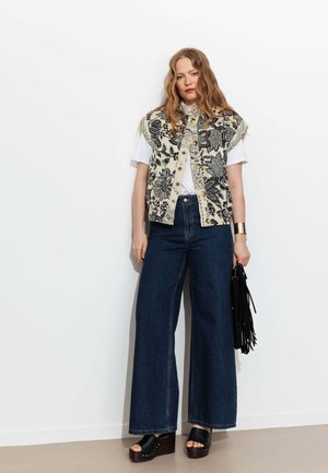 Femme aux cheveux ondulés portant un gilet fleuri sur un tee-shirt blanc, jean large foncé, sandales compensées noires, bracelet manchette doré, tenant un sac noir à franges.