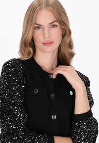 Fekete kardigán tüzdelő puff ujjakkal, ezüst gombokkal és zsebekkel, amely kiemeli az anyagok texturális kontrasztját.