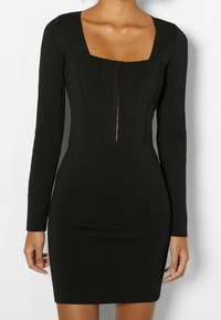 Robe noire ajustée avec manches longues, décolleté carré et détails de coutures verticales. Dotée d'une fermeture centrale avec des accents discrets en métal.