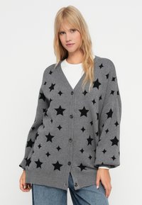 Grå cardigan med V-hals, svarta stjärnmönster, knäppning fram och neddragna ärmar, kombinerad med en vit skjorta och blåjeans.