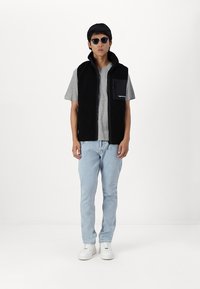 Zwarte fleece vest met hoge kraag en rits aan de voorkant, grijze t-shirt eronder, lichtgewassen jeans en witte sneakers. Accessoires zijn onder andere een zonnebril.