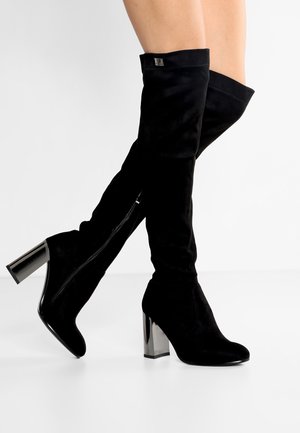 Musta polven korkeuteen ulottuvat saappaat, joissa on kiiltävät paksut korot, jalat ristissä ja vaaleaa taustaa vasten.