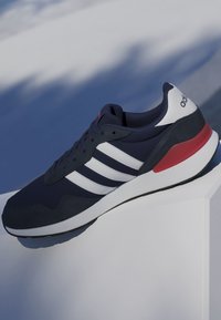 Zapatilla Adidas azul marino con rayas blancas y detalle rojo en el talón exhibida sobre una plataforma blanca sobre un fondo azul.