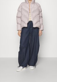 Puffig lavendelfärgad jacka med chevronmönster, hög krage och dragkedja; kombinerad med vida mörk denimjeans och vita sneakers.