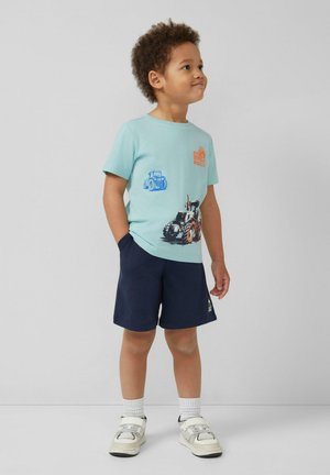 Garçon debout, les mains dans les poches, portant un t-shirt bleu clair avec un imprimé tracteur, un short bleu marine, des chaussettes blanches et des baskets sur fond uni.