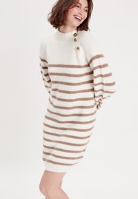 Robe pull crème avec des rayures horizontales beiges, dotée d'un col haut et de trois boutons dorés sur l'épaule gauche. Texture tricotée.
