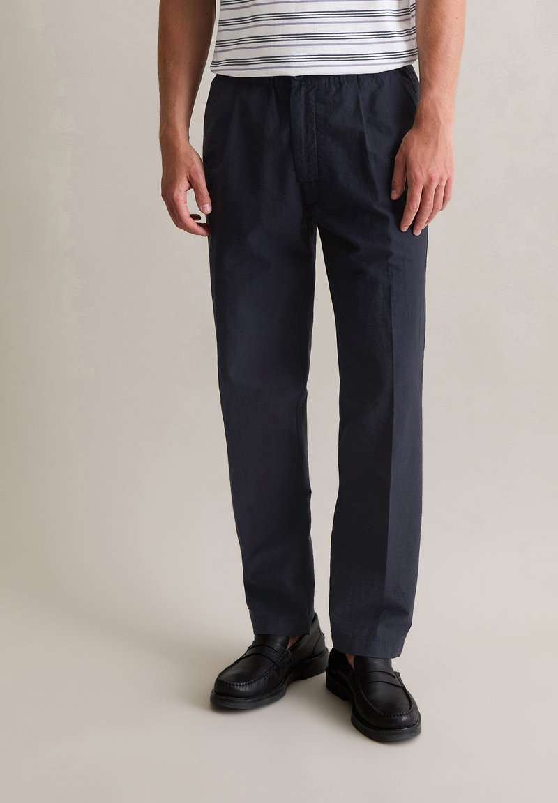 Marineblauwe, lichte broek met elastische taille en een ontspannen pasvorm. Geaccentueerd met subtiele textuur en gecombineerd met zwarte instappers.