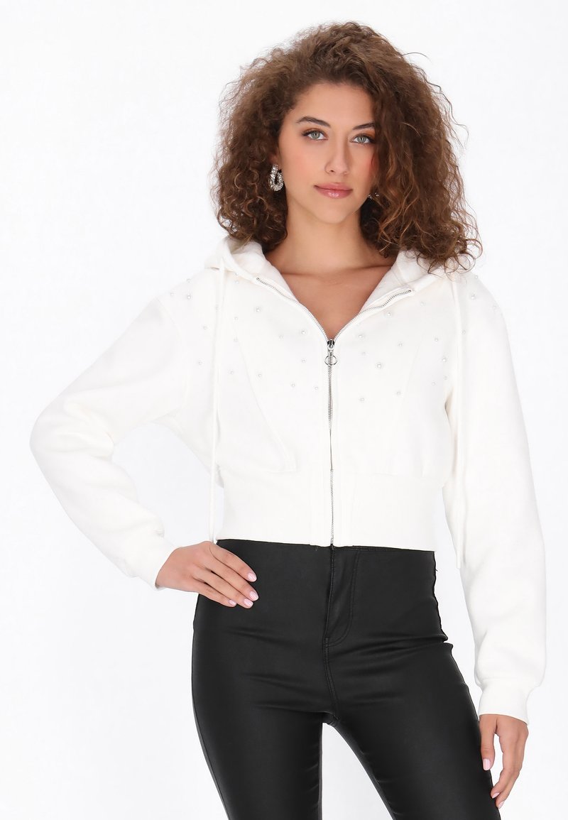 Hanorac alb scurt prevăzut cu fermoar frontal, decorat cu mici tăvițe. Are glugă cu șnur și mâneci lungi, asortat cu pantaloni negri.