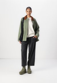 Jachetă verde cu guler maro, pulover bej, pantaloni largi gri închis și botine verzi. Modelul stă în picioare cu o geantă de umăr neagră.