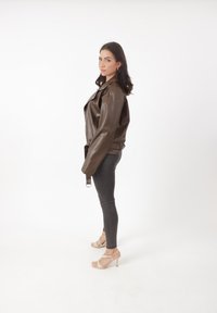 Veste en cuir marron à la coupe décontractée, avec de larges revers et un détail de ceinture sur le côté, associée à un jean gris ajusté et des sandales légères à brides.