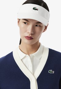 Visiera bianca con logo verde Lacoste, indossata con un cardigan blu navy caratterizzato da un colletto a coste bianche e dettagli con bottoni, design casual.