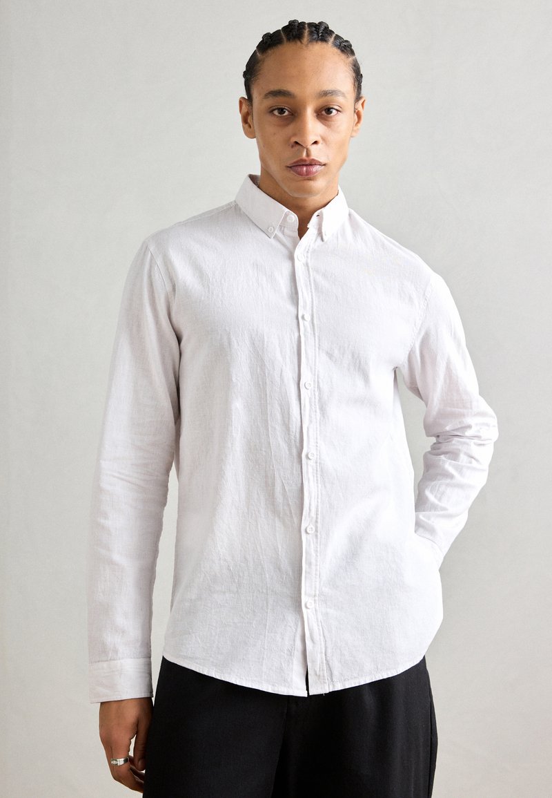 Camisa blanca de botones, hecha de un tejido texturizado, con cuello, mangas largas y dobladillo redondeado; combinada con pantalones negros.