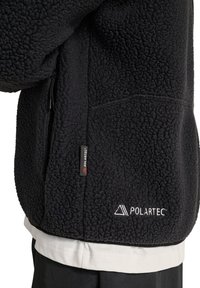Schwarze Polartec-Fleecejacke mit sichtbarer seitlicher Reißverschlusstasche und weißer Polartec-Logo-Stickerei nahe dem Saum.