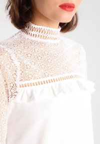 Blusa bianca con colletto in pizzo alto decorato con motivi floreali, dettagli arricciati lungo l'orlo e una texture di tessuto liscia e fluida.