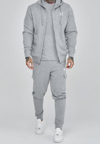 Grau zip-up Kapuzenpullover und passende Jogginghosen aus weichem Stoff, mit einem Logo auf der Brust und Cargo-Taschen an den Hosen, kombiniert mit wei�ßen Sneakers.