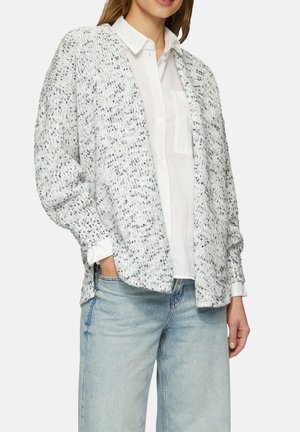 Gestructureerde witte cardigan met zwarte spikkels, open voorkant, verlaagde schouders en lange mouwen, gedragen over een witte overhemd met knopen en lichte spijkerbroek.