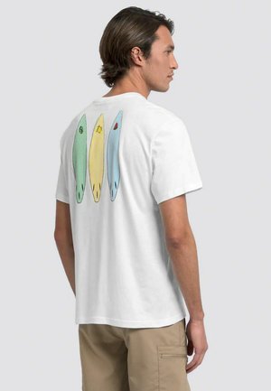 Hombre con camiseta blanca con ilustraciones de tablas de surf verdes, amarillas y azules en la espalda, combinado con pantalones cortos beige, de perfil.
