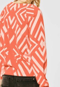 Coral long-sleeve top with white geometric patterns; soft texture, relaxed fit, round neck, and cuffed sleeves. Black jeans visible at the bottom.