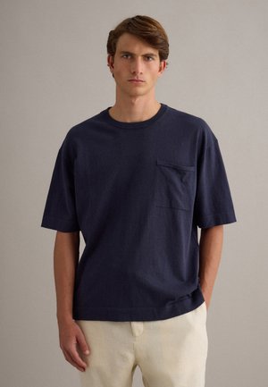 Giovane uomo con capelli castani corti che indossa una camicia blu navy scuro a maniche corte con taschino sul petto e pantaloni beige chiaro.