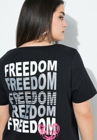Zwarte t-shirt met het woord "VRIJHEID" in verschillende vetgedrukte lettertypen en texturen, waaronder zilveren glitter en gestreepte ontwerpen, op de achterkant.