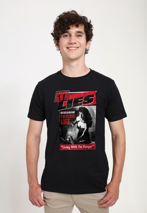 Ung mand med krøllet hår iført en sort t-shirt med en rød og hvid grafik med en kvinde og tekst om et hensynsløst liv.