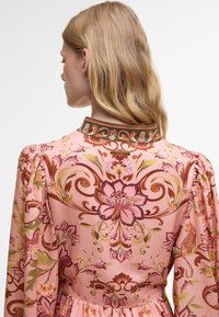 Robe rose à motifs floraux avec des manches longues, des épaules froncées et un col accentué de vert. Dotée de motifs complexes et de textures élégantes.
