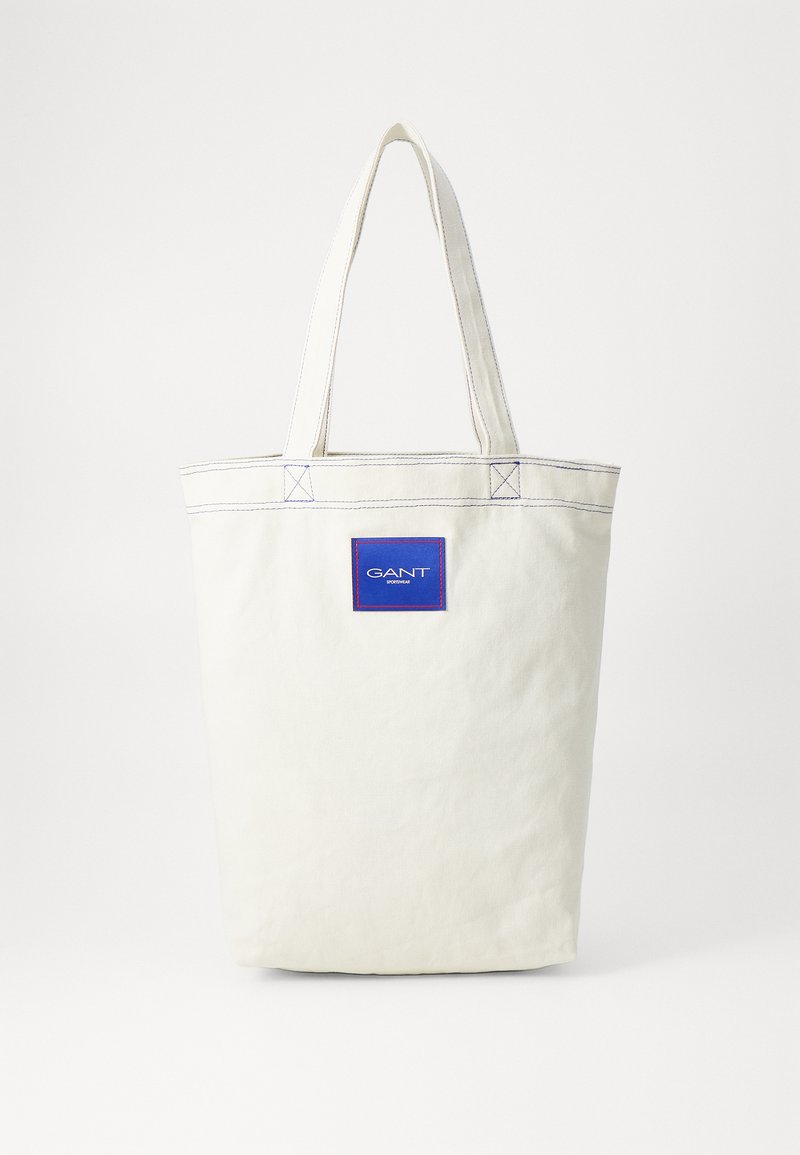 Weiße Stofftasche aus Leinen mit blauen Akzenten und Logo. Verfügt über robuste Griffe, verstärkte Nähte und eine einfache, rechteckige Form.