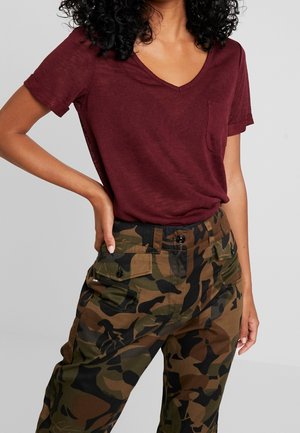 Woman wearing a burgundy V-neck T-shirt and camo cargo pants with buttoned pockets, one hand resting on her hip, against plain background.