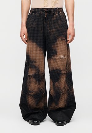 Brązowe i czarne szerokie spodnie tie-dye ze sznurkiem w pasie, przetarciami i czarnymi materiałowymi mankietami zakrywającymi czarne buty.
