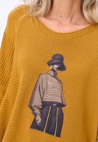 Musztardowożółty dziergany sweter o prążkowanej fakturze. Z ozdobnym nadrukiem przedstawiającym postać w pasiastym topie i kapeluszu typu bucket hat.