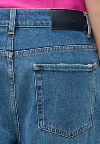 Prim-plan al buzunarului din spate al blugilor din denim albastru, cu o ușoară uzură, o etichetă de brand din piele neagră și un tricou roz băgat în pantaloni.