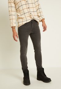 Calças jeans slim-fit pretas combinadas com uma camisa de botão bege e xadrez. A camisa apresenta riscas laranja e marinho num tecido texturizado. Botas pretas com atacadores.