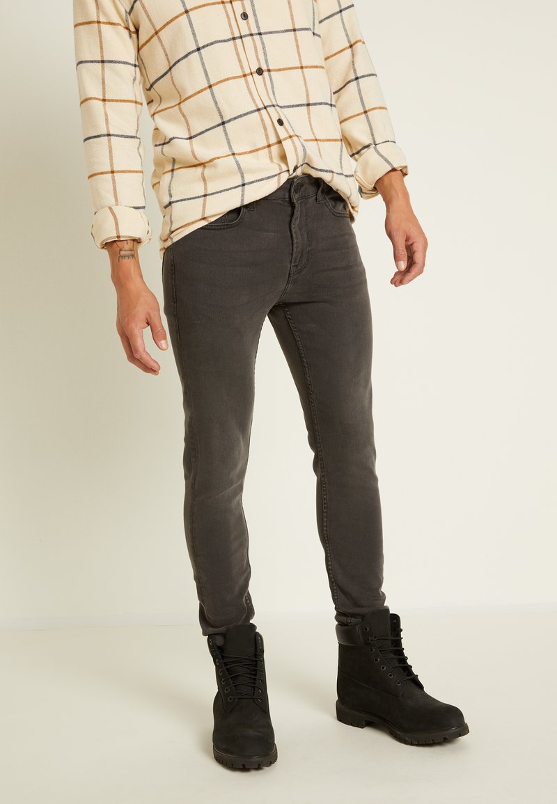 Calças jeans slim-fit pretas combinadas com uma camisa de botão bege e xadrez. A camisa apresenta riscas laranja e marinho num tecido texturizado. Botas pretas com atacadores.
