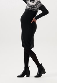 Svart stickad klänning med ett mönstrat tröjliknande överdel, kroppsnäv silhuett, knälängd. Bärs med svarta ankelskor och strumpbyxor.