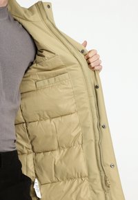 Una persona con un suéter gris sostiene abierta una chaqueta acolchada beige, mostrando el interior acolchado con un pequeño bolsillo interior.