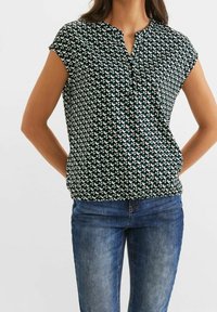 Krátký rukáv, zelená a bílá vzorovaná blůza s výstřihem a nařasením v pase, doplněná modrými džíny.