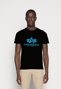 Schwarzes Baumwoll-T-Shirt mit einem blauen Logo, das ein stilisiertes "A" und Flügel-Elemente zeigt. Der Text lautet "ALPHA INDUSTRIES, KNOXVILLE, TENNESSEE, U.S.A."