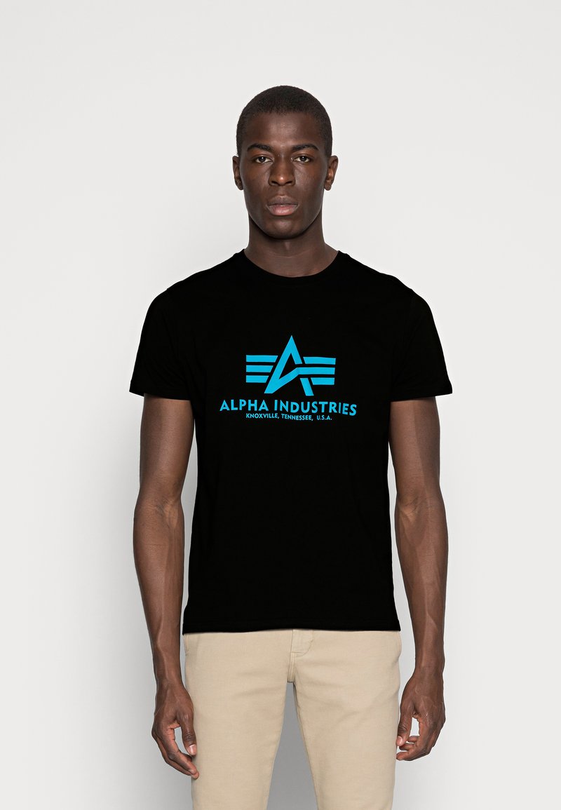 Schwarzes Baumwoll-T-Shirt mit einem blauen Logo, das ein stilisiertes "A" und Flügel-Elemente zeigt. Der Text lautet "ALPHA INDUSTRIES, KNOXVILLE, TENNESSEE, U.S.A."