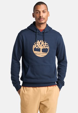 Donkerblauwe hoodie van zachte stof, met een groot tan boomlogo, een voorzak en een capuchon met trekkoord. Afgemaakt met ribgebreide manchetten.