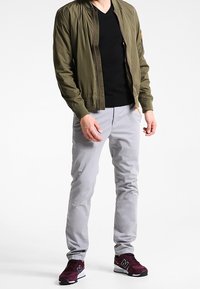 Olivgrön bomberjacka, svart V-ringad topp, ljusgrå slim-fit byxor och vinröda sneakers med vita detaljer.