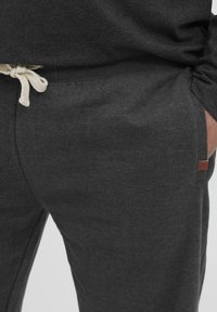 Un pantalon de survêtement gris foncé en tissu doux, doté d'un cordon de serrage de couleur crème contrastante et d'une petite étiquette de logo marron sur le côté.