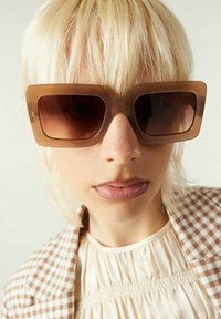 Personne blonde portant de grandes lunettes de soleil carrées brunes, un haut smocké beige et une veste à carreaux bruns et blancs.