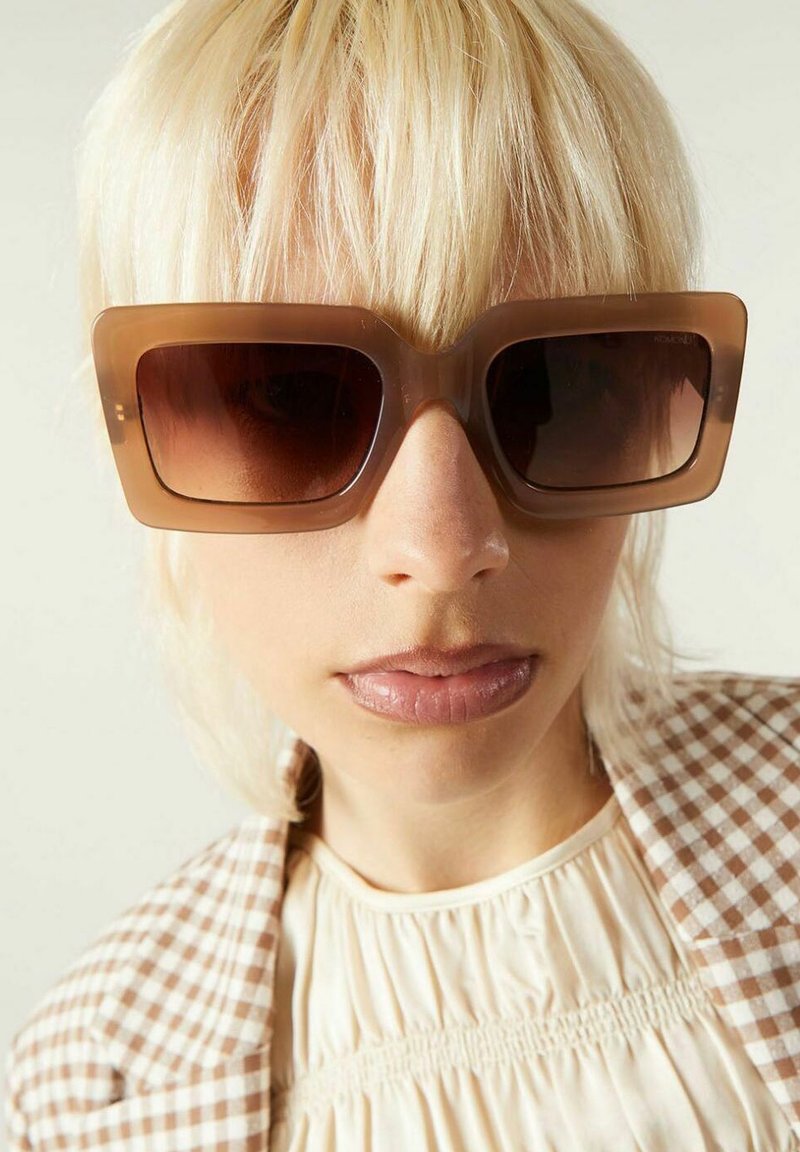 Personne blonde portant de grandes lunettes de soleil carrées brunes, un haut smocké beige et une veste à carreaux bruns et blancs.