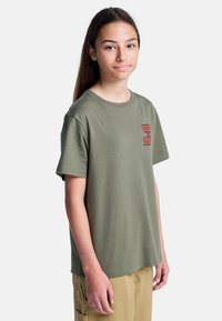 Camiseta de algodón verde oliva con un corte relajado, mangas cortas y un estampado gráfico rojo elevado en el área del pecho izquierdo. Textura suave.