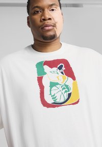 Tricou alb cu un grafic colorat al unei pisici stilizate care ține o minge de baschet, cu accente verzi, roșii și galbene în jurul designului.